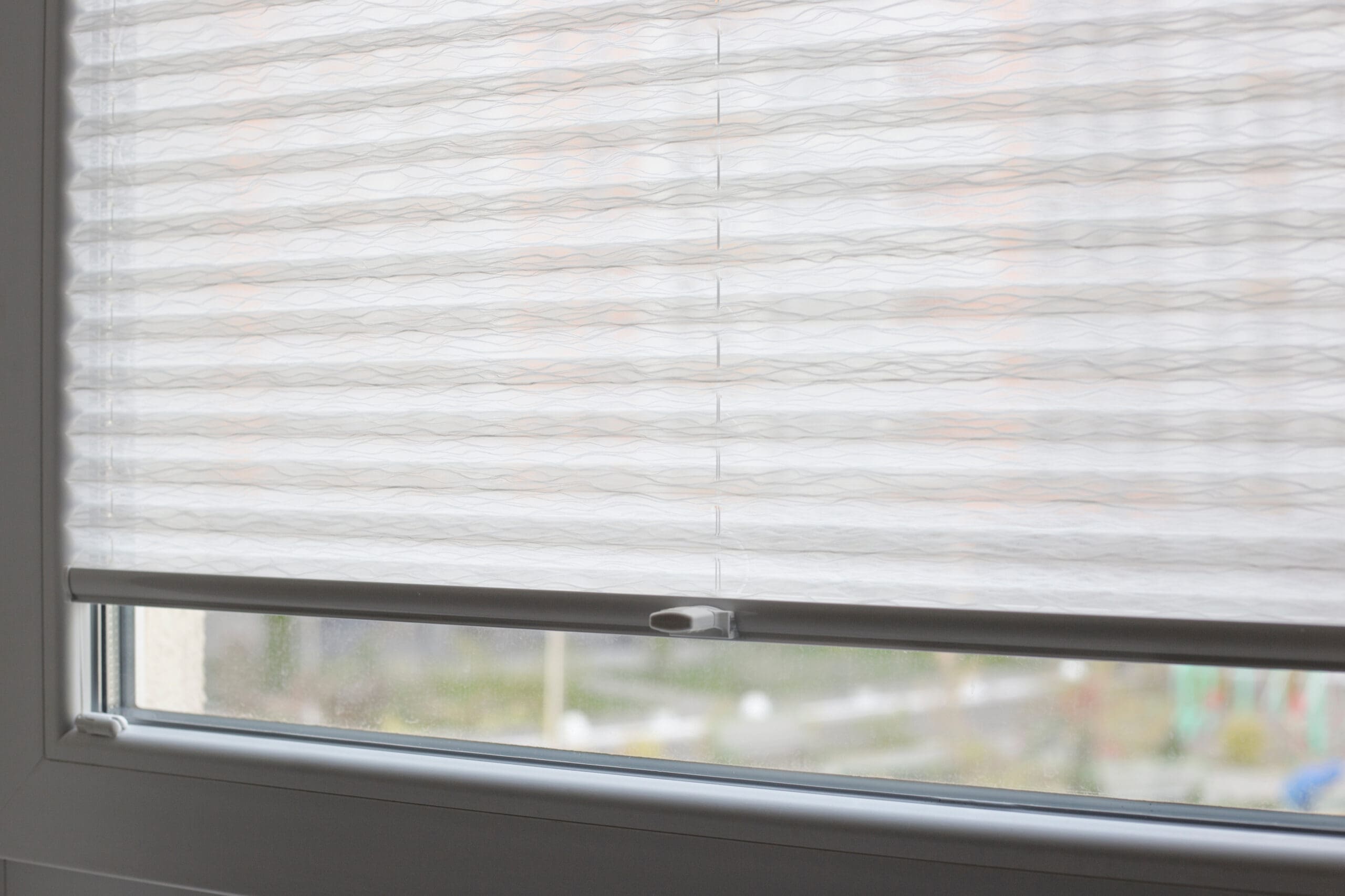 A close-up view of a window with white, wavy-patterned made to measure blinds partially closed, allowing diffused sunlight to filter through and obscuring the view of the outside.