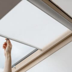 A hand pulls down a white, made to measure blind over a skylight window set in a light wooden frame.