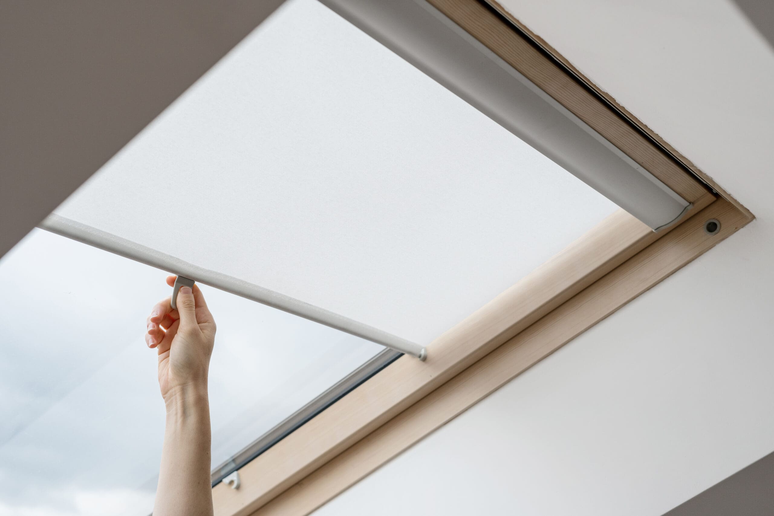A hand pulls down a white, made to measure blind over a skylight window set in a light wooden frame.