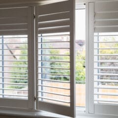 An open window with white Wooden Shutters, partially opened to reveal a view of houses, greenery, and a wooden fence outside.