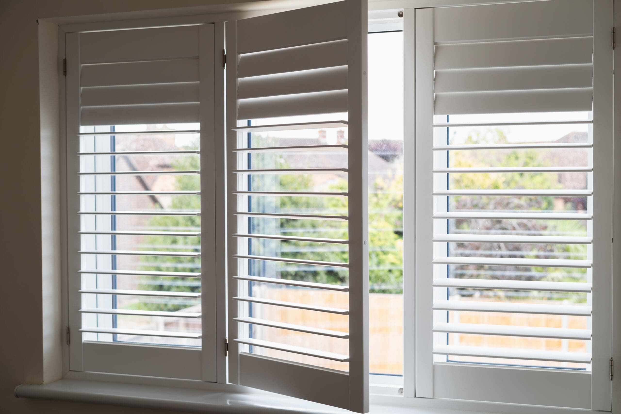 An open window with white Wooden Shutters, partially opened to reveal a view of houses, greenery, and a wooden fence outside.
