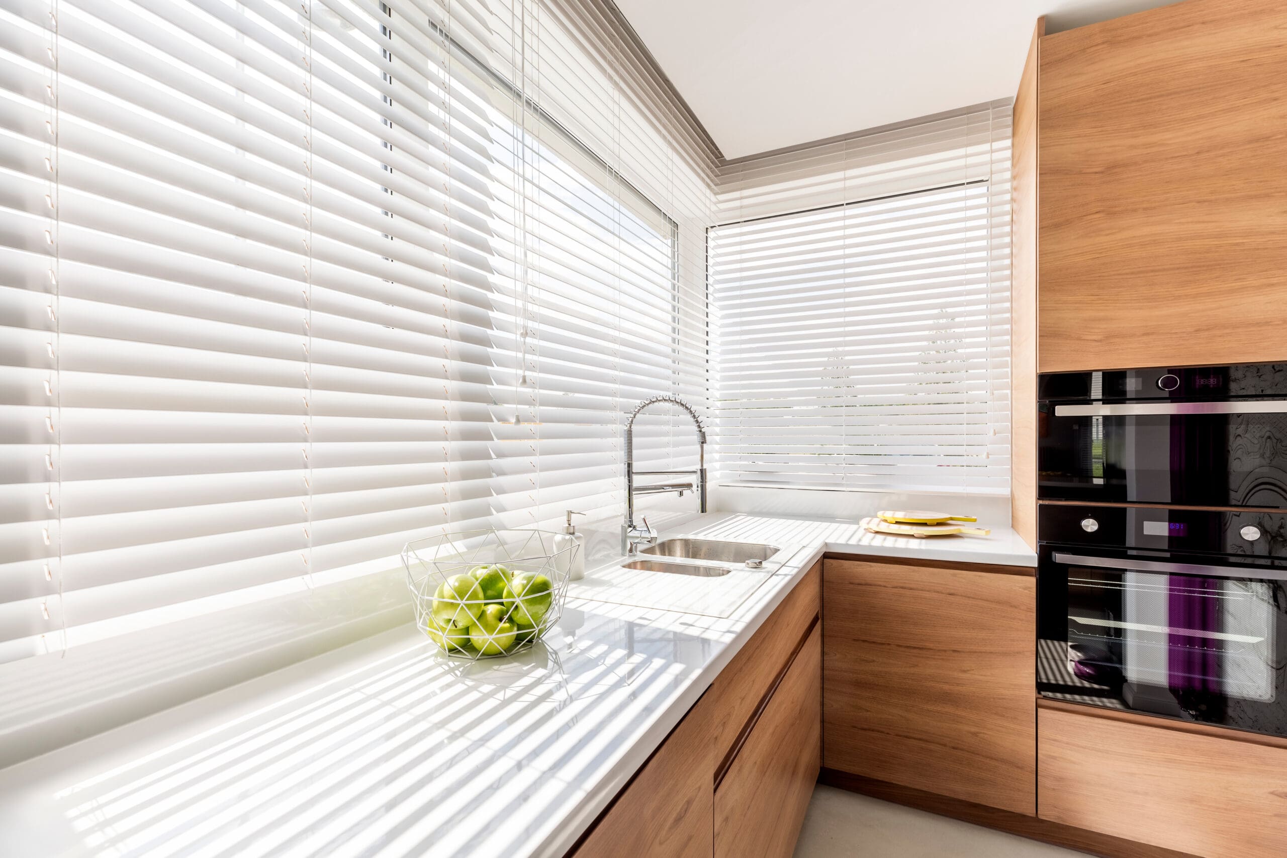 Modern kitchen with light wood cabinets, white countertops, a stainless steel sink, a bowl of green apples, and large windows with made to measure blinds letting in sunlight. A built-in black oven is visible on the right.