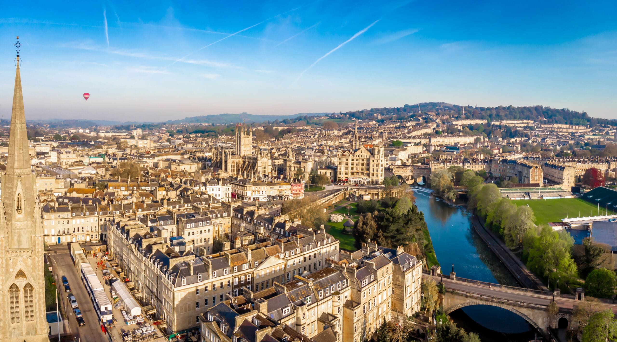 Aerial view of Bath, England, showing historic stone buildings with conservatory blinds, a church spire, the River Avon, green hills in the background, and a hot air balloon floating in the clear blue sky.