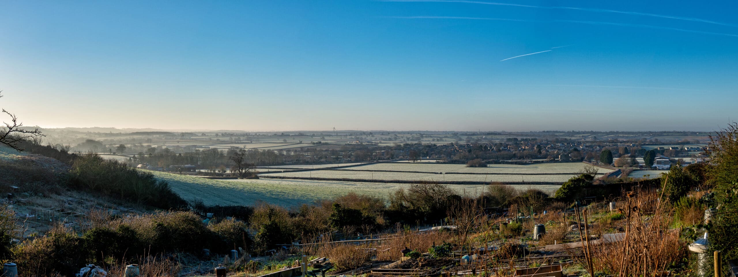 Panoramic view of a frosty rural landscape with fields, hedgerows, and a small village featuring homes fitted with Perfect-Fit Blinds under a clear blue sky on a bright, cold morning.