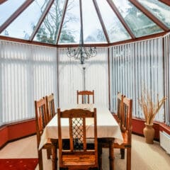 A sunlit conservatory dining area with a glass ceiling, wooden table and six chairs, white tablecloth, chandelier, decorative vase with branches, and stylish Conservatory Blinds offers beautiful views of the garden outside.