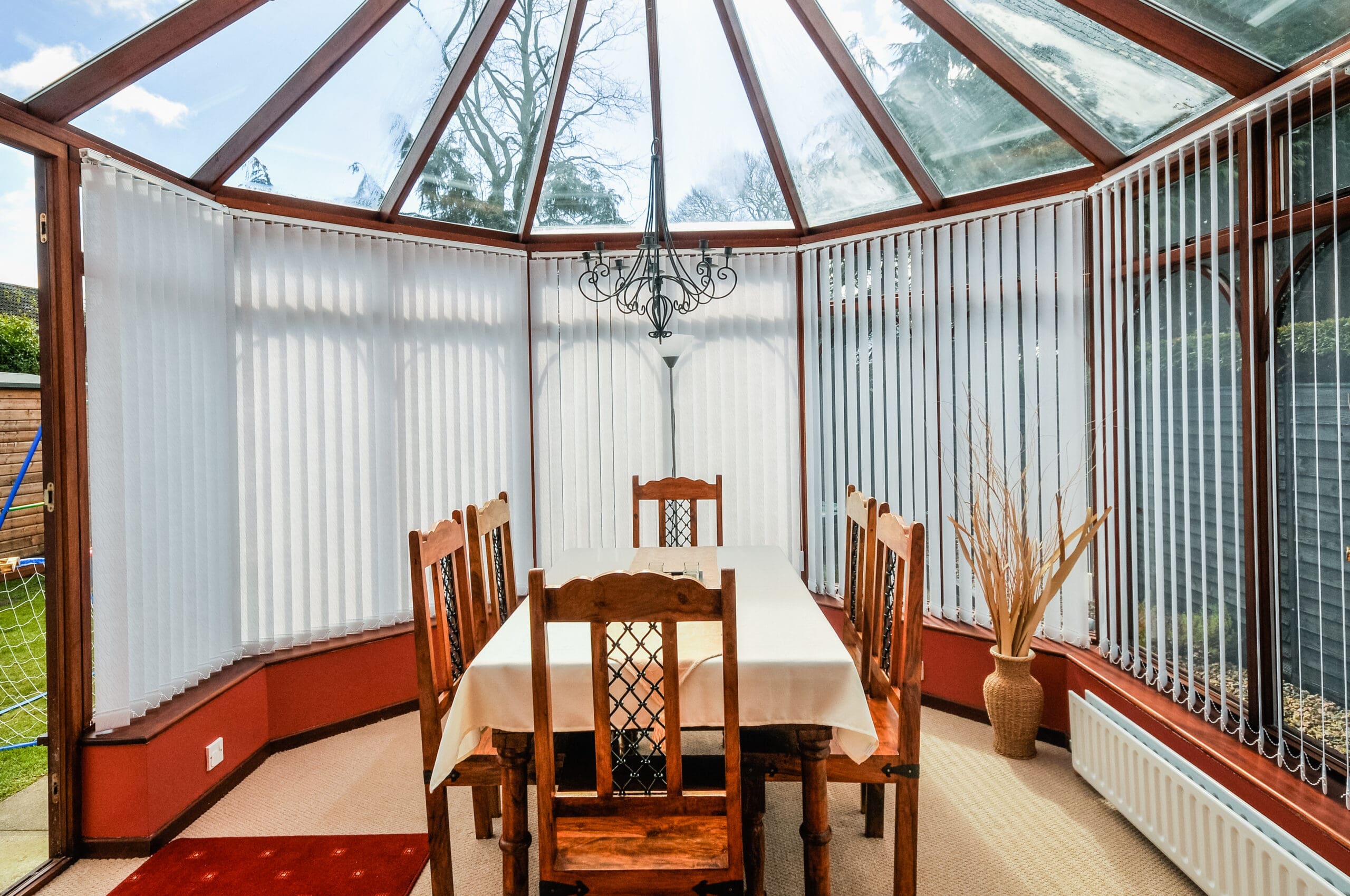 A sunlit conservatory dining area with a glass ceiling, wooden table and six chairs, white tablecloth, chandelier, decorative vase with branches, and stylish Conservatory Blinds offers beautiful views of the garden outside.