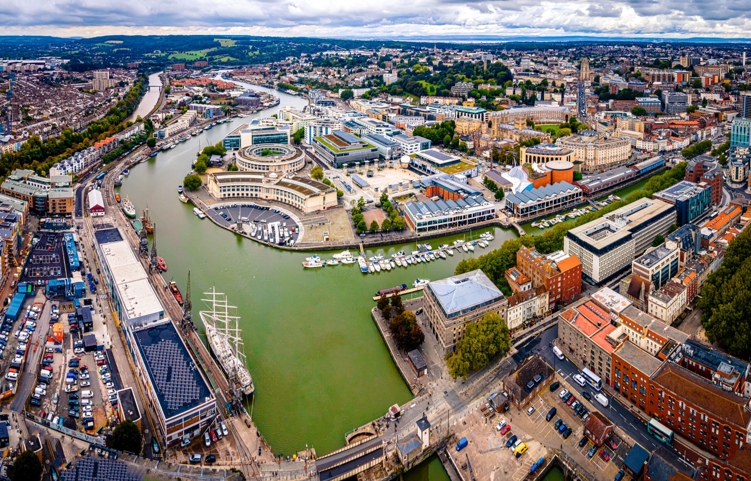 Aerial view of a city with a river running through it, modern and historic buildings, a docked tall ship, boats in the marina, and green hills beyond—some homes visible feature elegant wooden shutters under the cloudy sky.