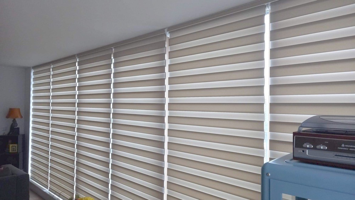 A row of large windows covered with beige made to measure blinds allows some light to filter through. There is a record player and a blue shelf in the foreground, and a lamp in the background.