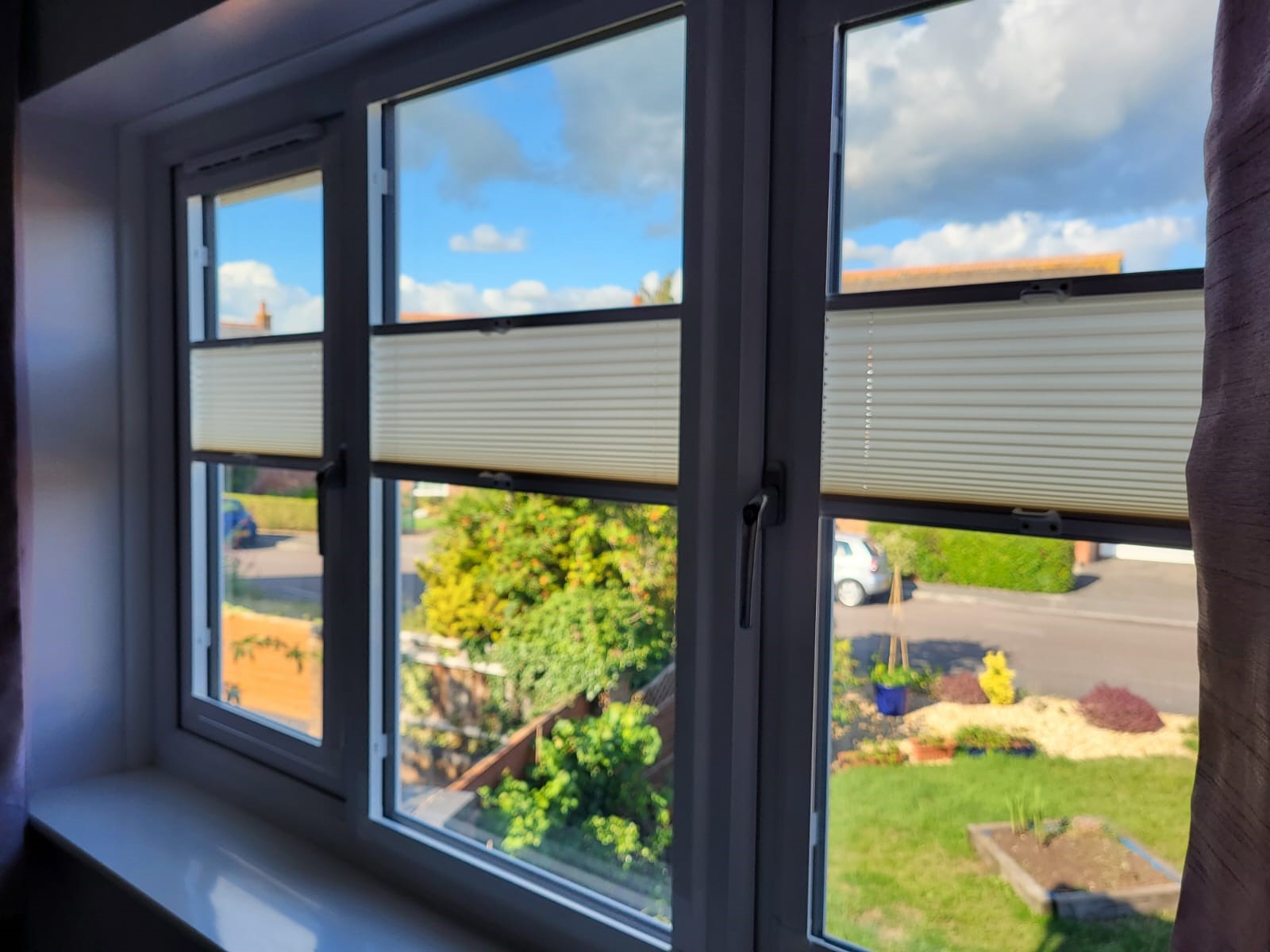 A view through a window with partially closed made to measure blinds, showing a residential street, green bushes, a white car, and a blue sky with clouds outside.