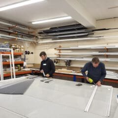 Two people work at a large table assembling window frames in a Blind Shop workshop. Shelves are filled with tools, supplies, and long pieces of metal or plastic. Bright ceiling lights illuminate the workspace.