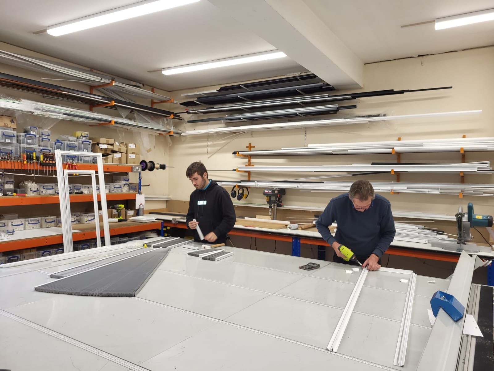 Two people work at a large table assembling window frames in a Blind Shop workshop. Shelves are filled with tools, supplies, and long pieces of metal or plastic. Bright ceiling lights illuminate the workspace.