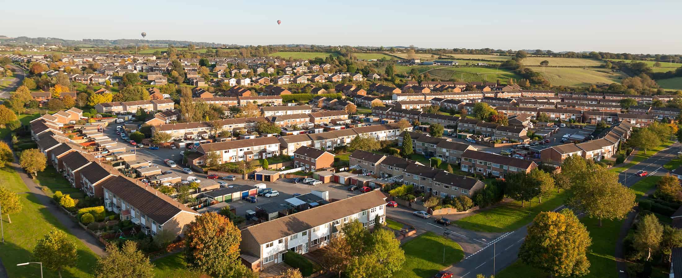 Aerial view of a suburban neighborhood with rows of houses, driveways, green trees, and surrounding fields. Roads curve around the houses, and hot air balloons are visible in the distant sky.